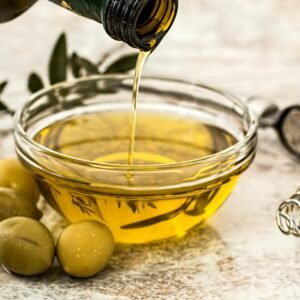 Close-up of olive oil being poured into a glass bowl surrounded by fresh olives and kitchen tools.